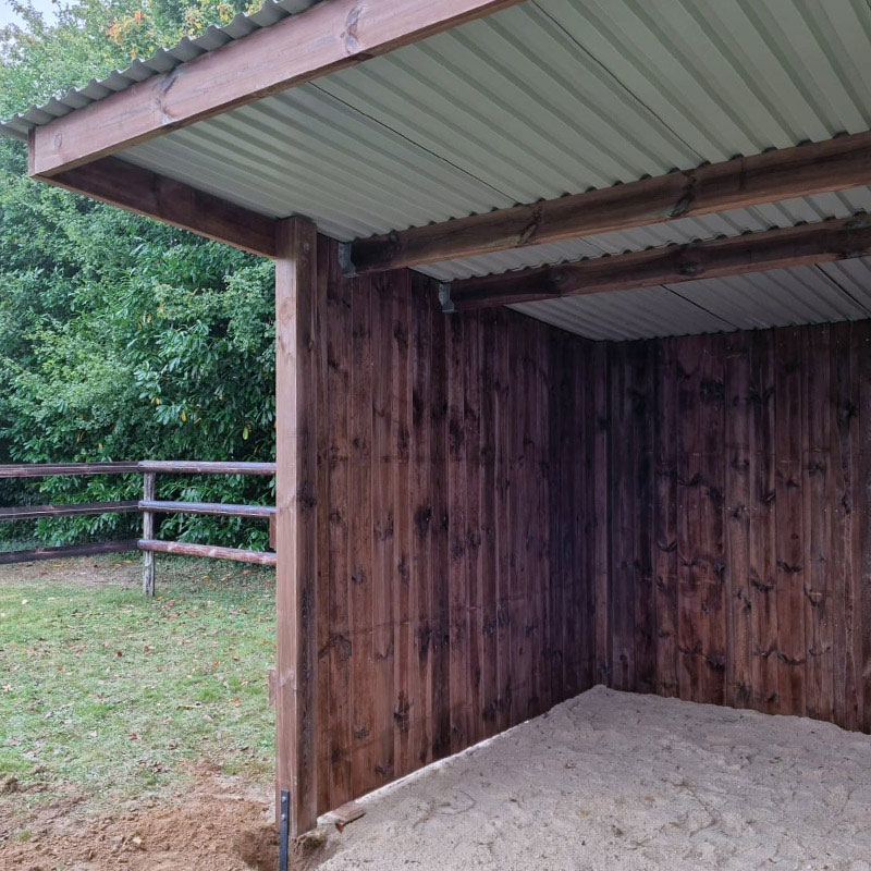 Abri pour chevaux en madrier, vue de l'intérieur, avec une barrière à 3 lisses en arrière-plan.
